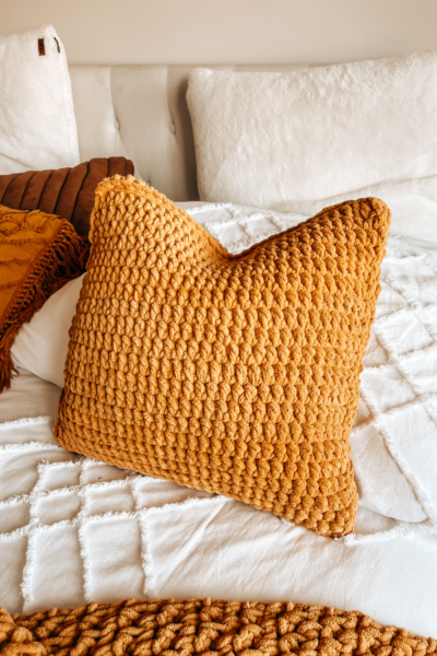 Una imagen de un cojin tejido a crochet de color mostaza sobre una cama blanca