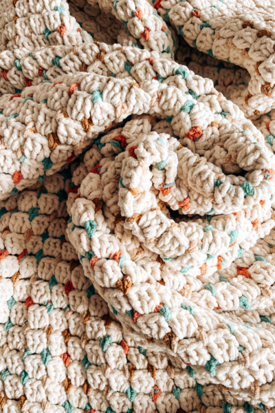 Imagen de una manta o cobija tejida a crochet en color crema con puntos de colores. La manta está retorcida en e medio