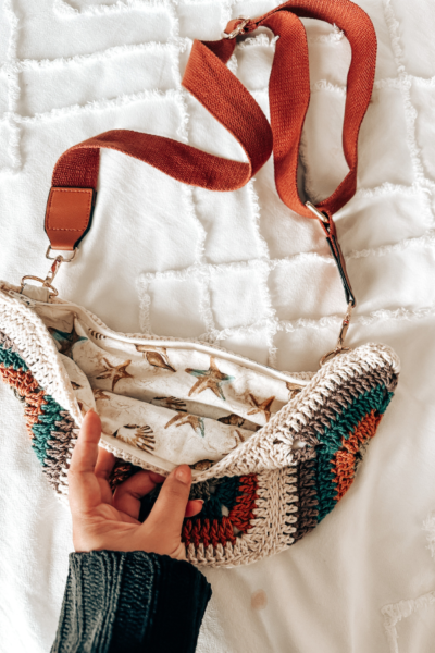 Un bolso tejido a crochet con granny squares con asas de tela cafe sobre una cama blanca.